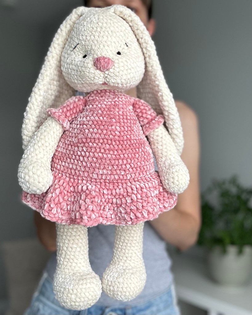 A crocheted bunny rabbit plush toy with a pink dress, showcasing soft yarn and detailed features, held by a person in a light-colored room.